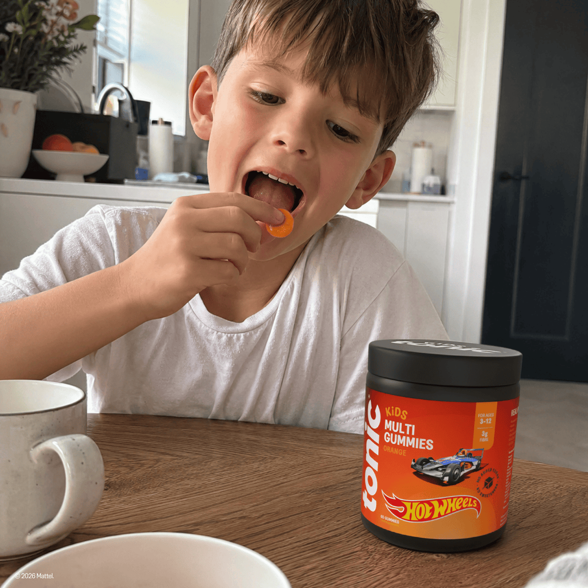 A young boy in a white t-shirt sits at a wooden table, opening his mouth to eat an orange Tonic Kids Multi Gummy. In the foreground, a tub of Tonic Hot Wheels Multi Gummies is prominently displayed on the table.