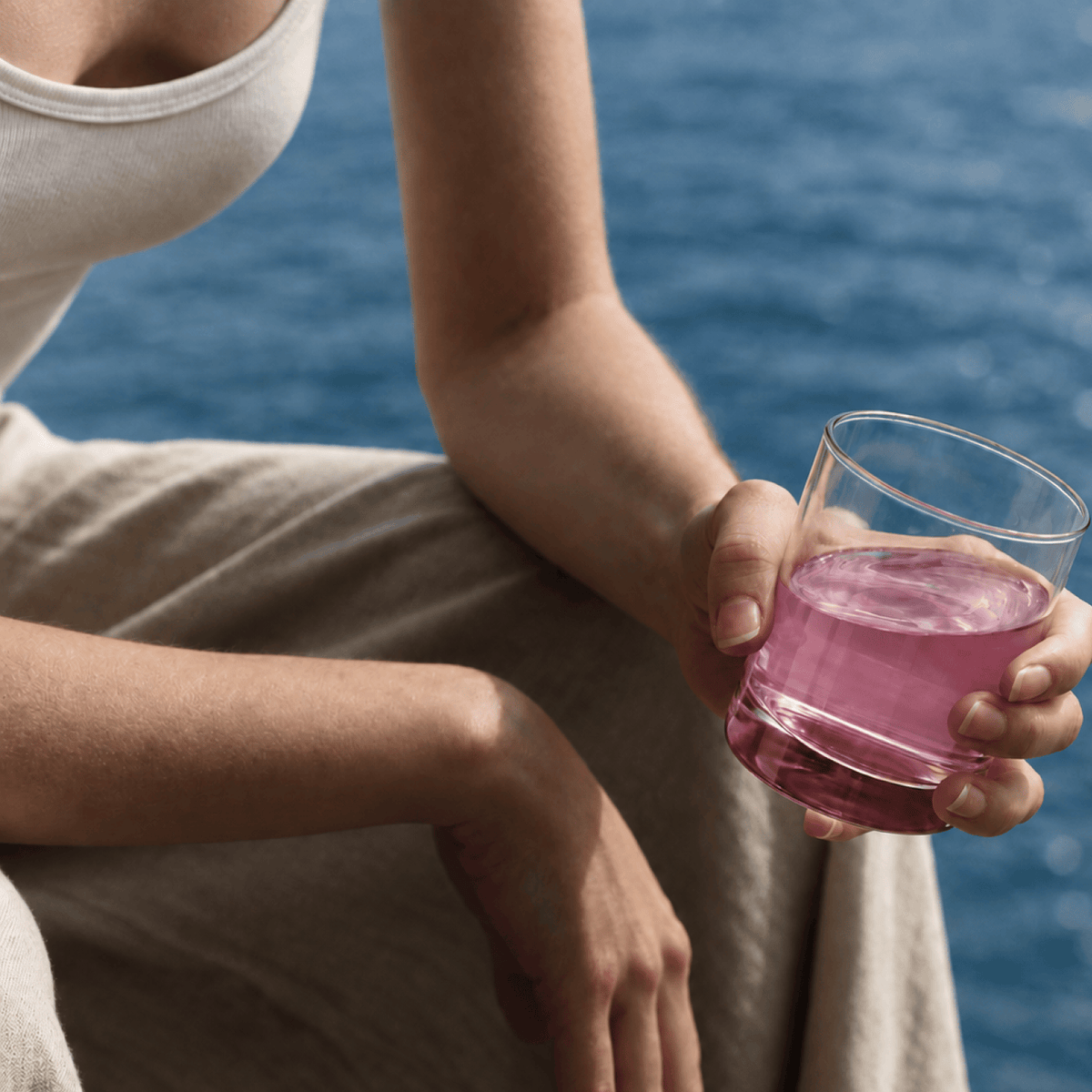 A person sitting outdoors by the blue water holds a glass of Tonic Creatine + Brain. The person is wearing a white tank top and beige trousers.