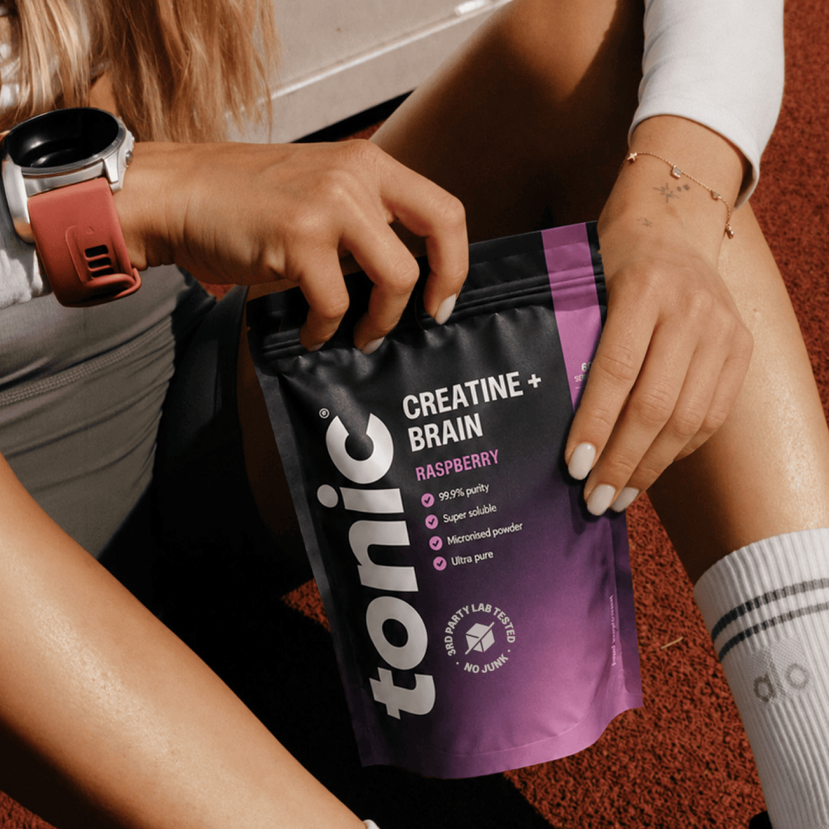 A person holding a black and purple Tonic Creatine + Brain Raspberry powder pouch, with their legs and hands visible against a red running track background.