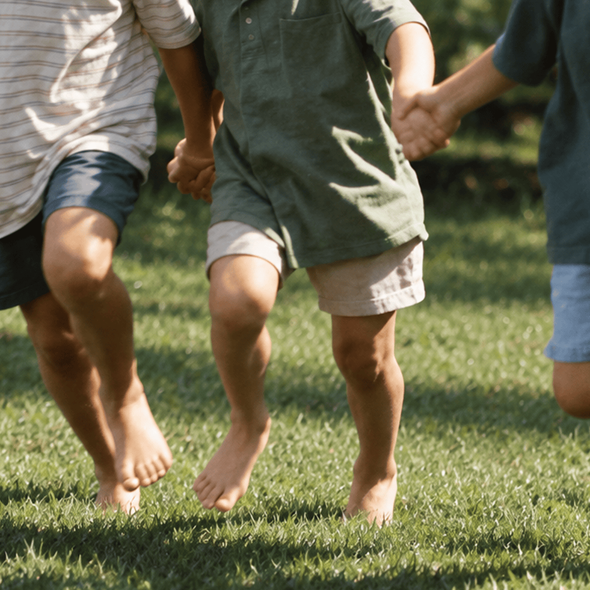 Three children running barefoot on a grassy lawn, holding hands. The center child wears a muted green short-sleeve shirt and light beige shorts, while other children in striped and blue shirts are partially visible.