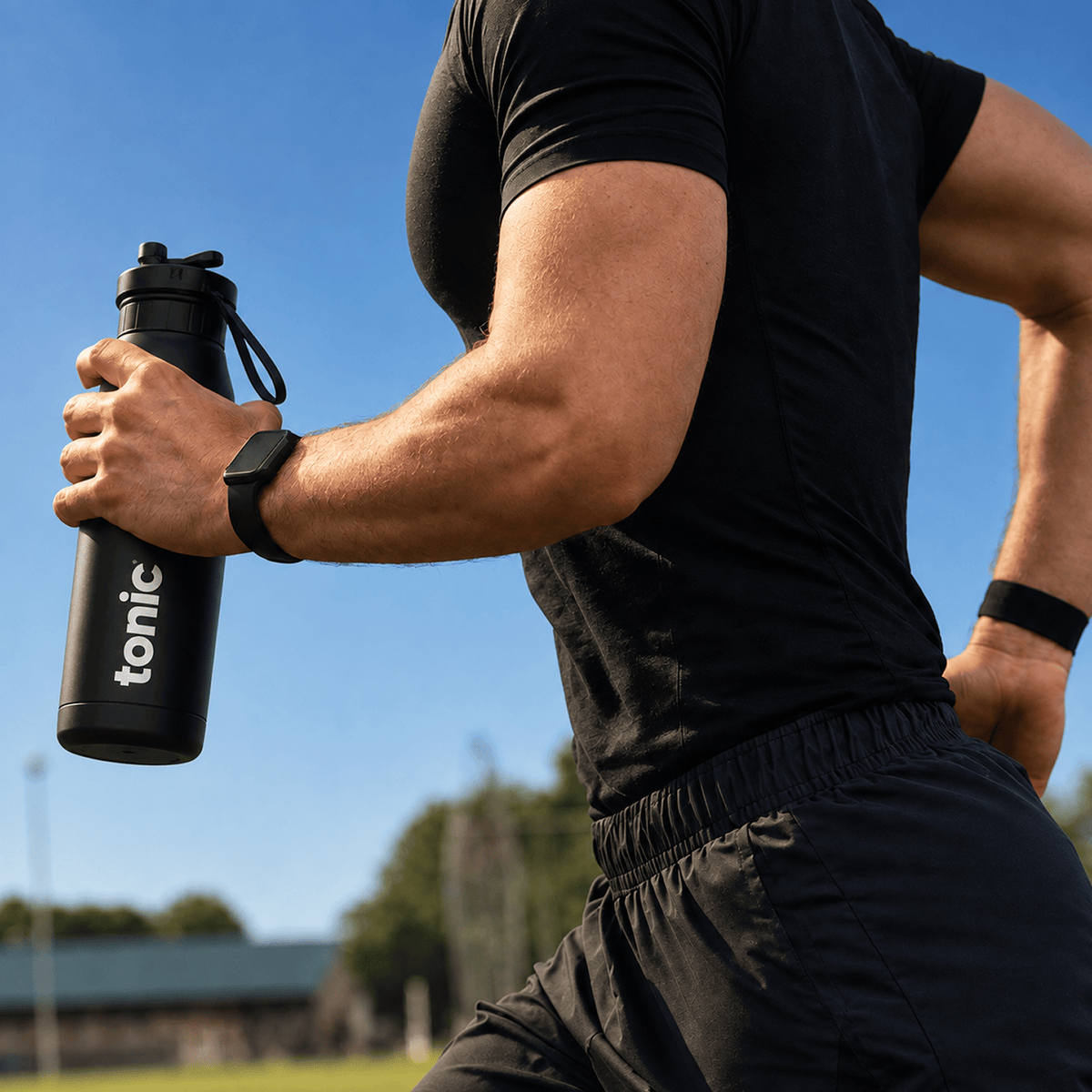 A close-up lifestyle shot of a man running outdoors, holding a black Tonic water bottle. He is wearing a black athletic t-shirt, black running shorts, a black smartwatch, and a black wristband, with a blue sky and green trees in the background.