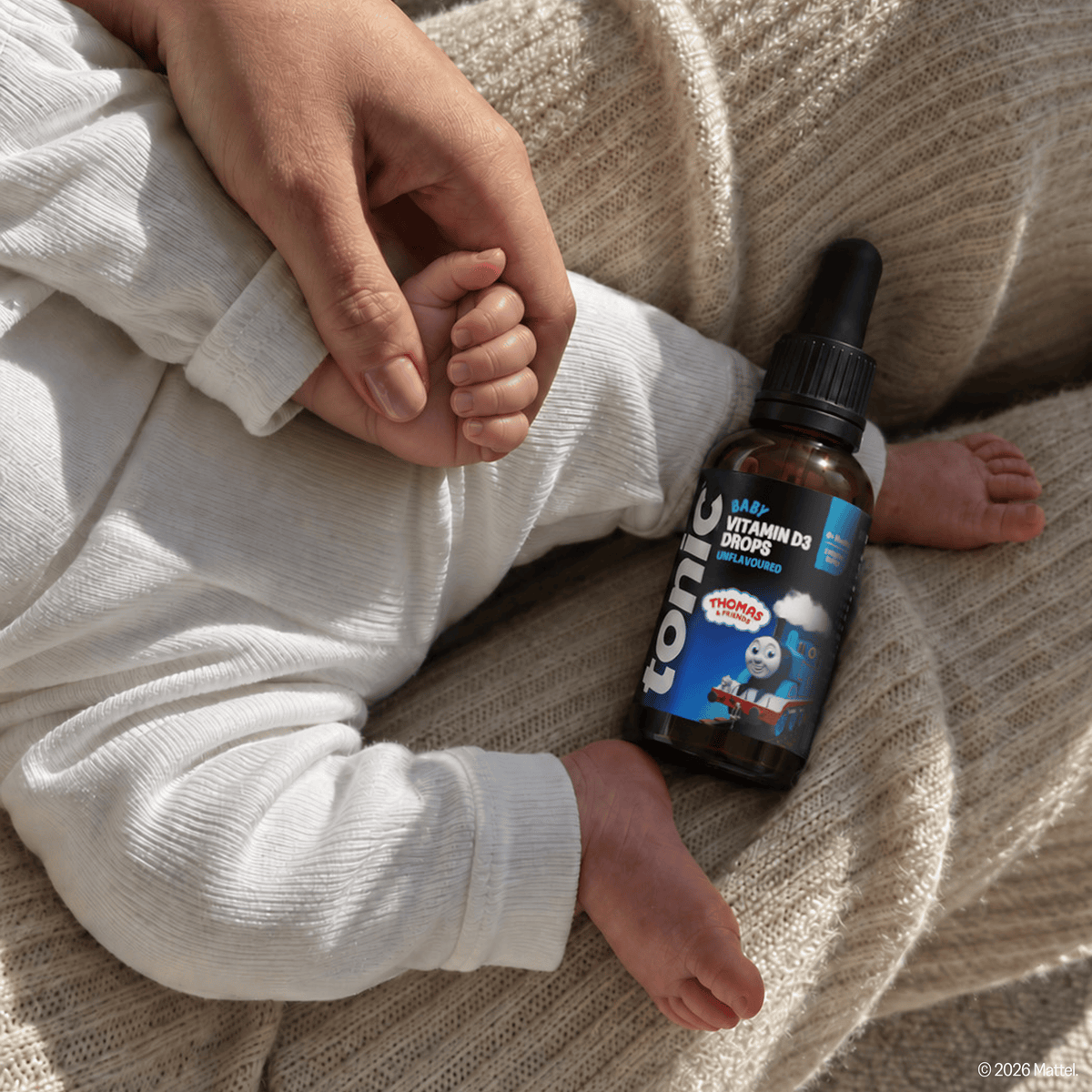 A baby in a white ribbed long-sleeve outfit rests on a beige knit blanket, holding an adult's hand. The baby's foot and arm are visible next to a small amber dropper bottle labeled 'Tonic Baby Vitamin D3 Drops'.