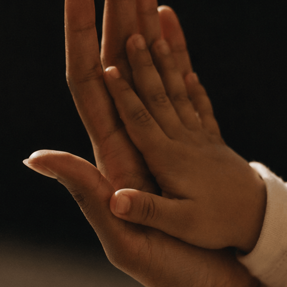 A close-up shot of two hands, one adult and one child, gently touching in a warm, intimate pose against a dark background.