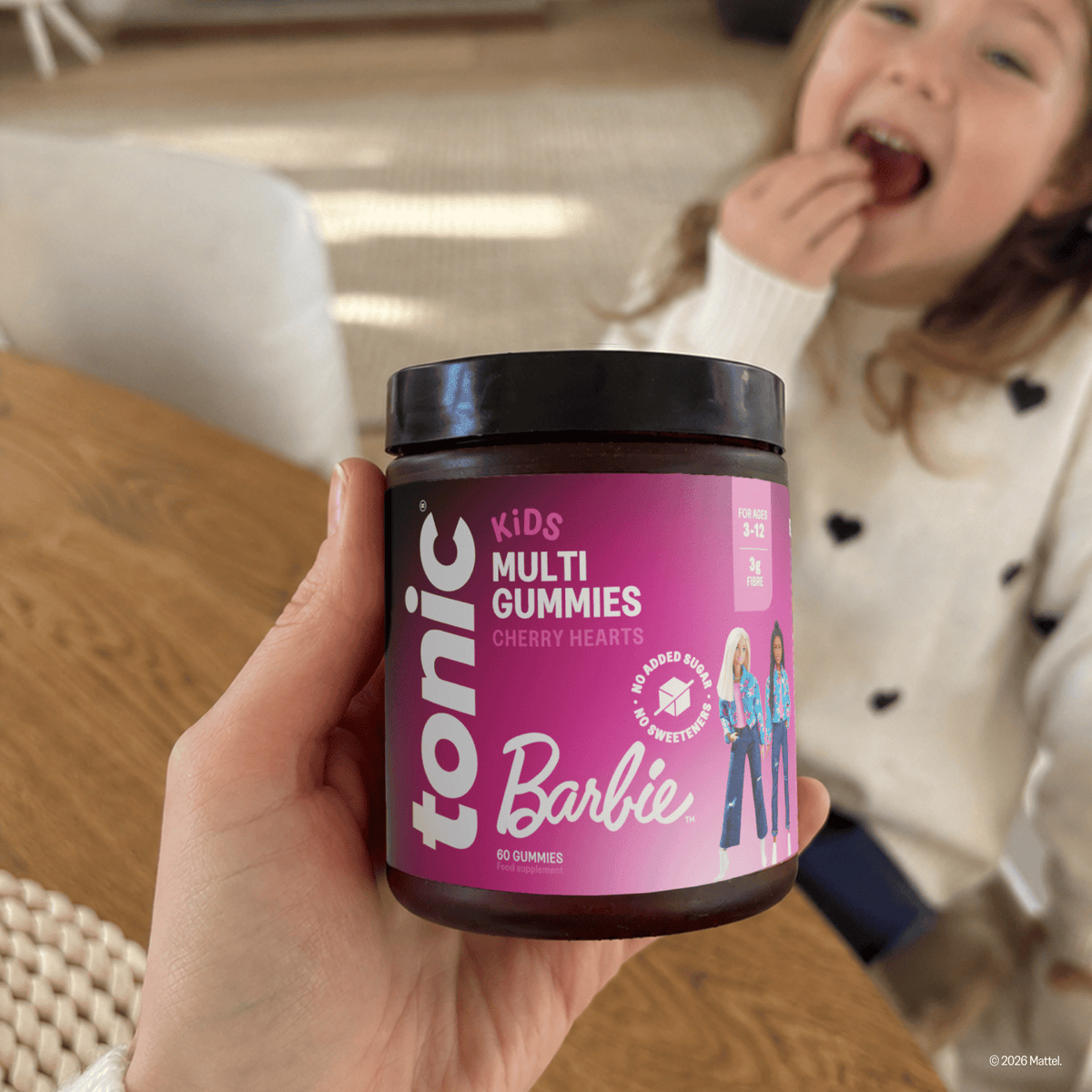 A hand holds a jar of Tonic Kids Multi Gummies in front of a young child eating in a indoor setting. The jar features a bright pink label with the Barbie logo and character illustrations.