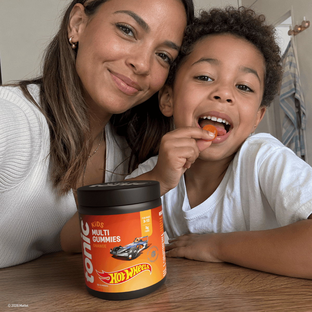 A mum and her son sit at a wooden table indoors, with the boy eating a gummy from a jar labeled 'tonic' and 'Hot Wheels Kids Multi Gummies'.