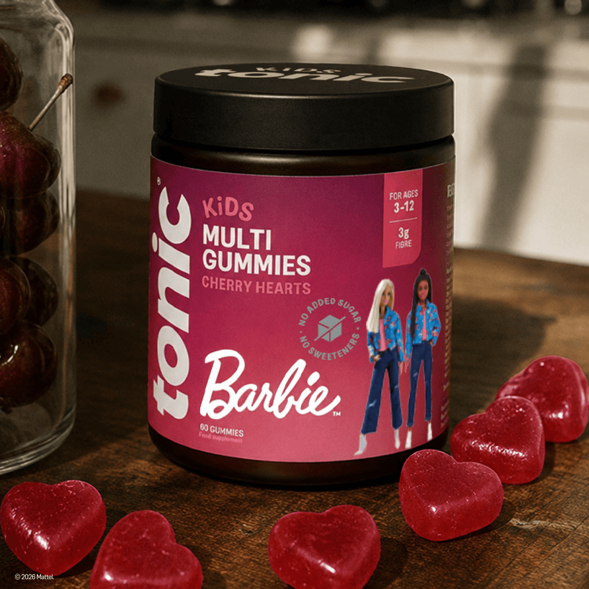 A jar of Tonic Kids Multi Gummies Cherry Hearts candy sits on a wooden table, surrounded by red heart-shaped gummies. A glass jar of cherries is visible on the left, and the background shows a softly blurred indoor setting.