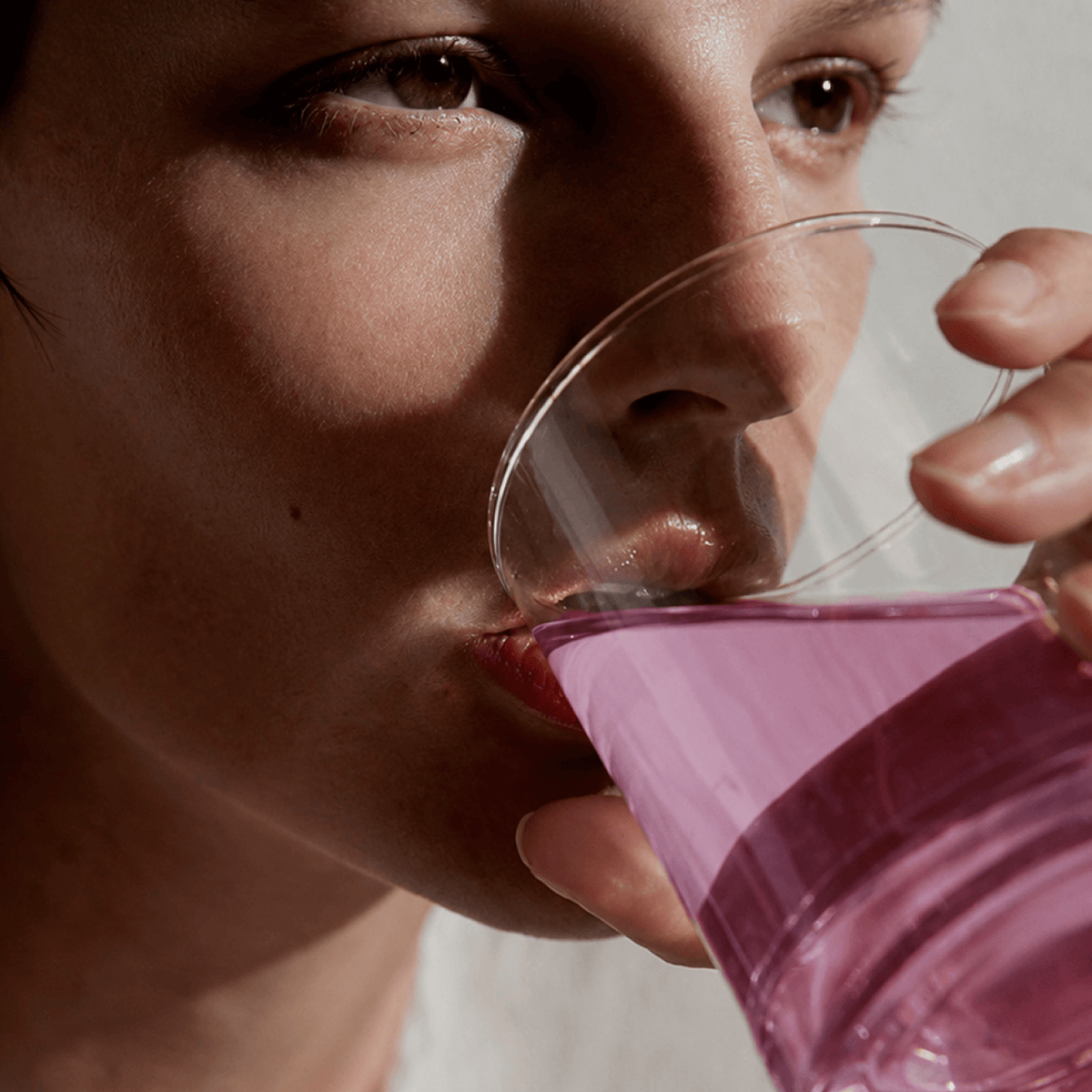 A close-up lifestyle shot of a person drinking from a clear glass containing Tonic Creatine + Brain with strong directional sunlight casting shadows on their face.