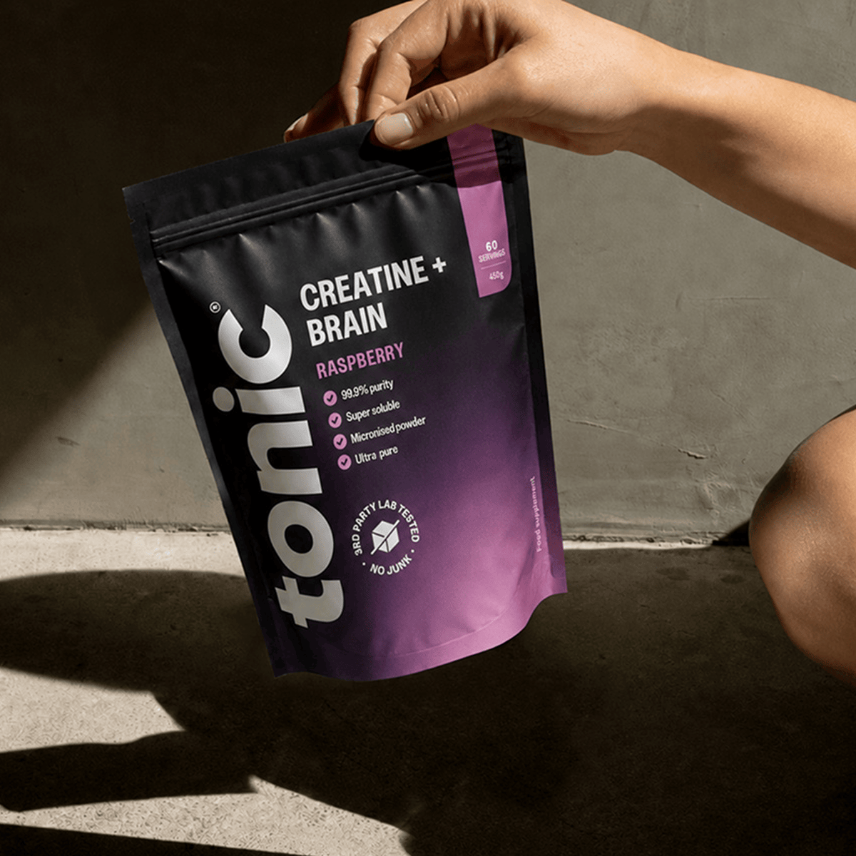 A woman holding a pouch of Tonic Creatine + Brain by the top, with a neutral background and shadows. 