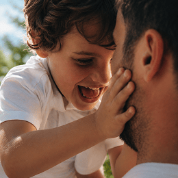A close-up lifestyle shot of a young boy with curly brown hair smiling and touching his father's face, with the father wearing a white t-shirt. The background shows a soft-focus outdoor setting with green foliage and blue sky.
