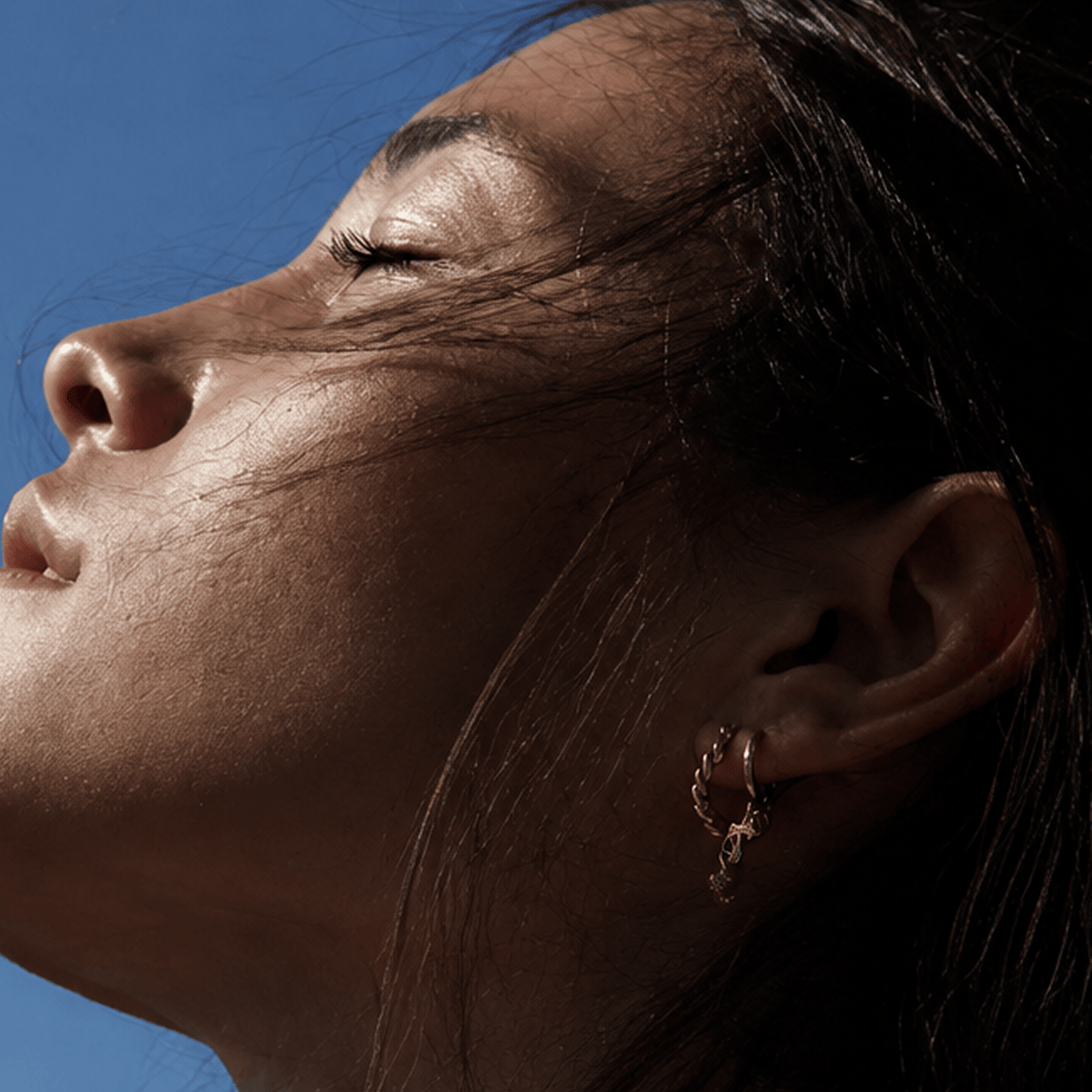 A close-up lifestyle shot of a woman's face with her head tilted back against a clear blue sky, wearing a small gold hoop earring.