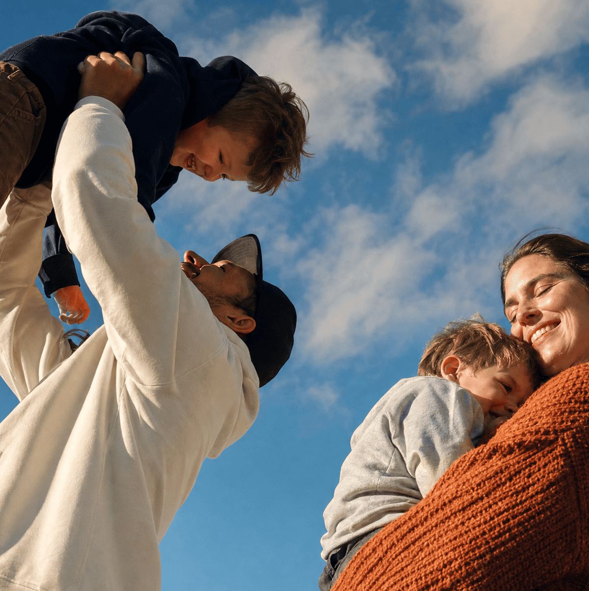 A family of four outdoors under a blue sky with clouds, captured from a low angle. A man in a cream sweatshirt and black cap lifts a young boy into the air, while a woman in a rust-orange knit sweater holds a sleeping toddler.