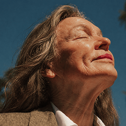 A close-up portrait of a woman with her eyes closed, looking upward, wearing a brown textured blazer over a white collared shirt against a blue sky background.