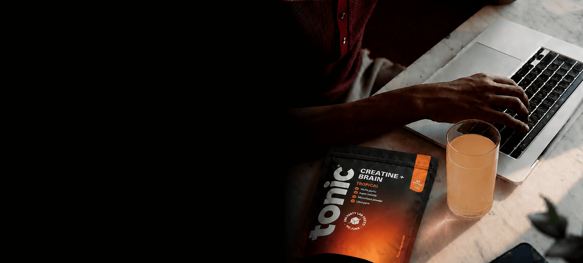 A person typing on a laptop at a desk with their hand visible, alongside a glass and pouch of Tonic Creatine + Brain on a marble surface.