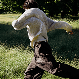 A person outdoors in a grassy field, wearing a cream open-knit sweater over a light shirt and dark brown pants, captured mid-motion.
