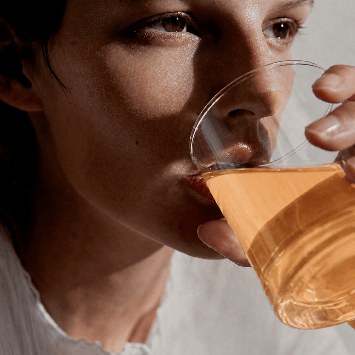 A close-up lifestyle shot of a person drinking from a clear glass containing Tonic Creatine + Brain, tropical flavour, with dramatic side lighting and a soft-focus neutral background.