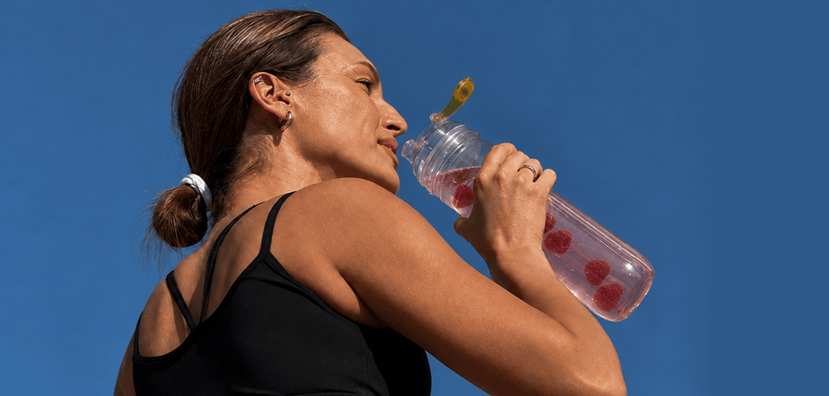 A woman in a black athletic tank top drinking Tonic Creatine + Brain from a bottle, set against a solid blue sky.