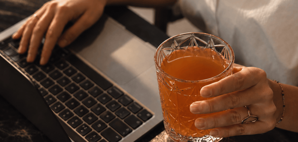 A close-up lifestyle shot of a person holding a glass Tonic Creatine + Brain while typing on a laptop. The person's hands and arms are visible, along with a white shirt and a dark desk surface.