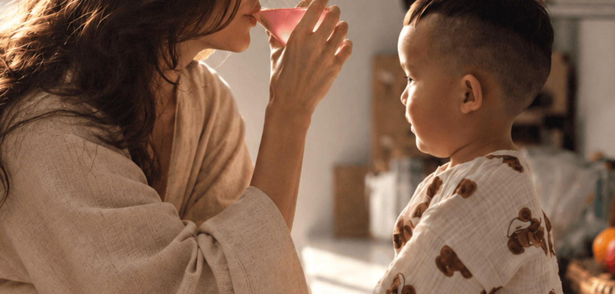 A woman in a beige linen robe drinks Tonic Creatine + Brain while a young child in a white linen shirt with a brown bear pattern looks on, set in a warm, softly blurred home interior.