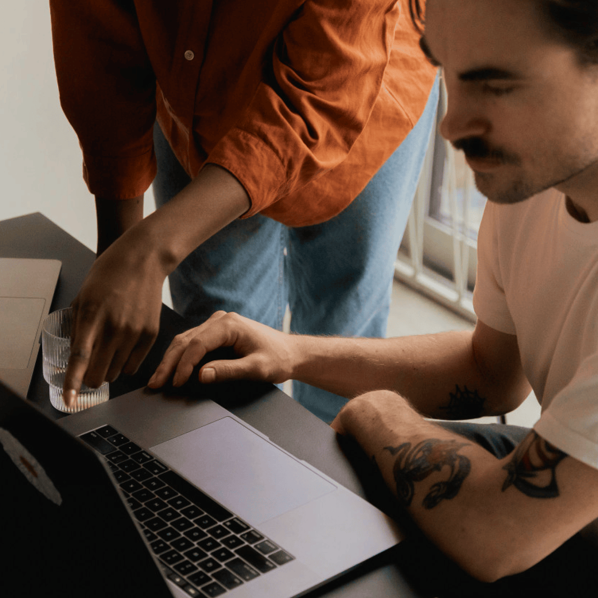 Two people collaborating at a desk with a laptop. One person in an orange shirt leans in while the other, wearing a white t-shirt.