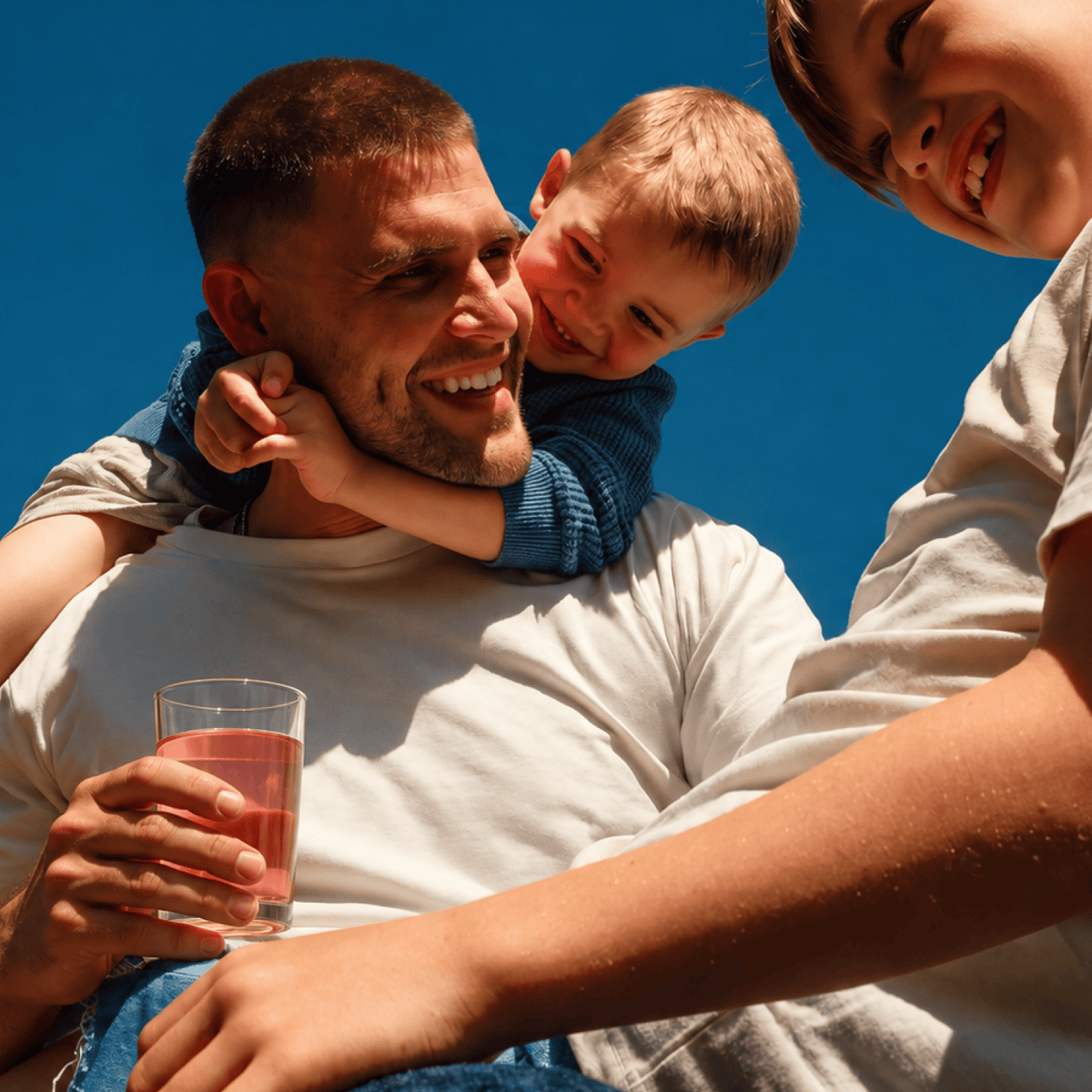 A smiling man in a white t-shirt holds a glass of Tonic Creatine + Brain while with two children against a solid blue sky. One child is on his shoulder and another is cropped on the right.
