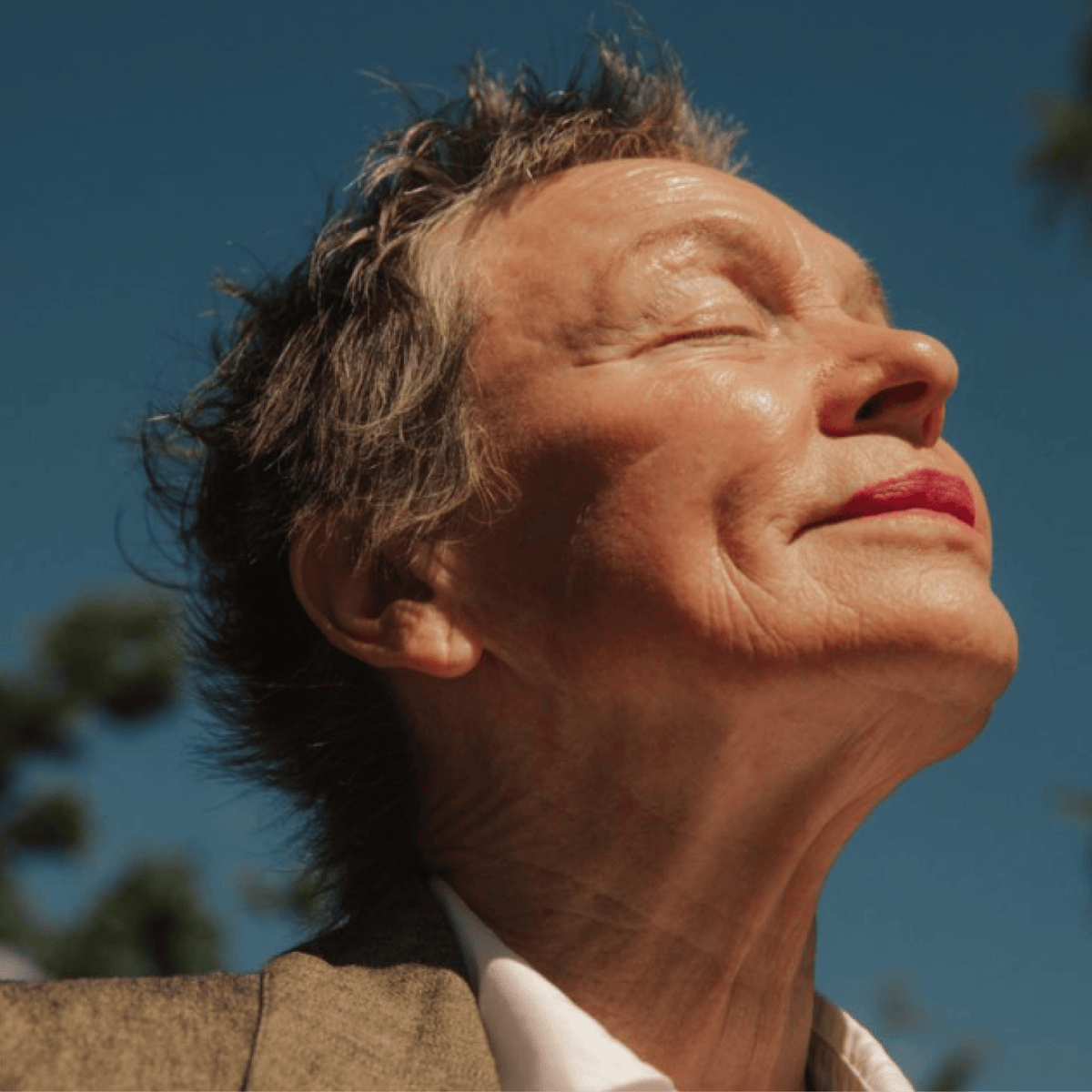 Close-up outdoor portrait of an older woman with her eyes closed and head tilted back, wearing a brown blazer and white collared shirt against a blue sky.