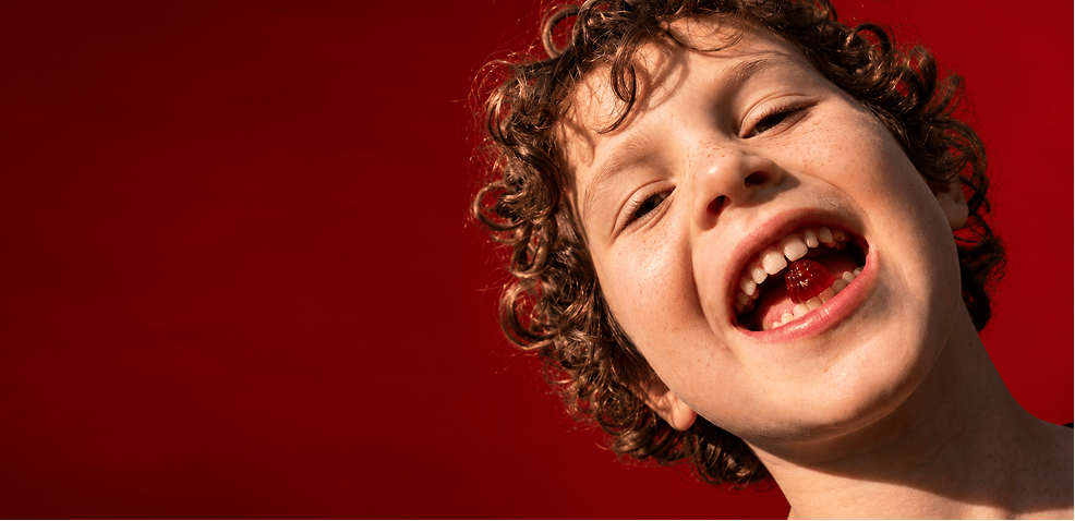 Laughing child with curly hair showing a strawberry Tonic Kids Multi Gummy held between their teeth, photographed against a bold red background