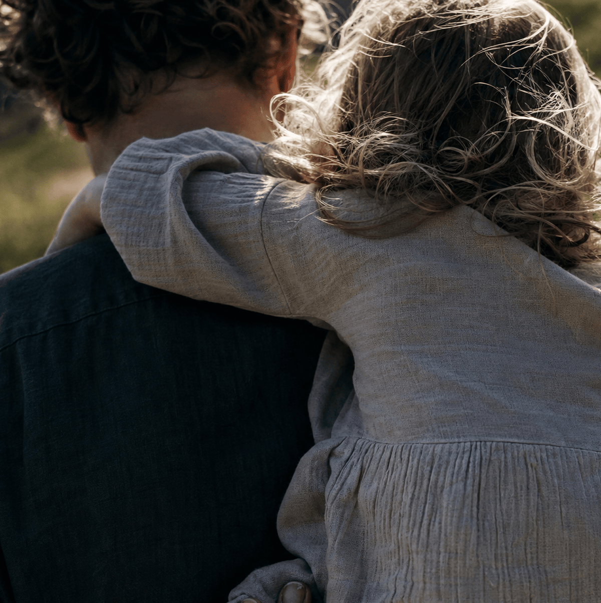 A close-up outdoor shot of a child wearing a light beige linen long-sleeve dress, leaning over an adult's shoulder. The child's hair is blowing in the wind, and the background is a soft, blurred natural green and gold bokeh.