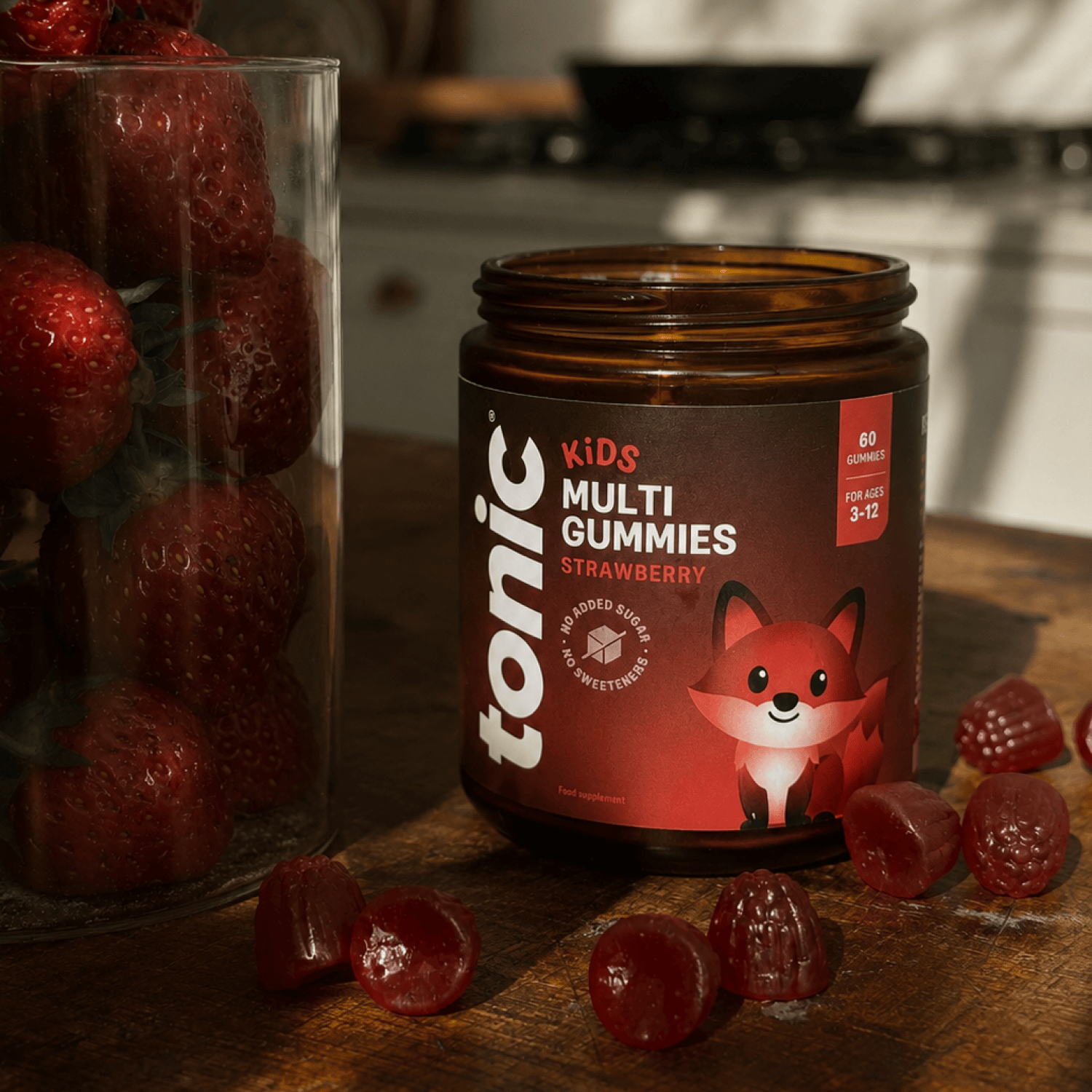 A jar of Tonic Kids Multi Gummies Strawberry is displayed on a wooden table next to a glass of fresh strawberries, with several red gummy candies scattered in front. The background shows a softly blurred kitchen setting.