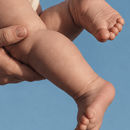 A close-up shot of a baby's bare legs and feet being held by an adult's hand against a solid blue background.